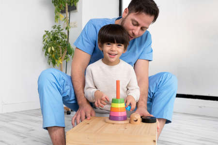 Young therapist helping cute little boy who has cerebral palsy, playing with developing toy at rehabilitation clinic.の写真素材