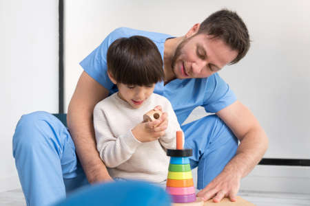 Young therapist helping cute little boy who has cerebral palsy, playing with developing toy at rehabilitation clinic.の写真素材