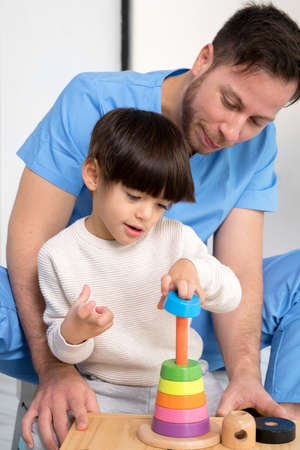 Young therapist helping cute little boy who has cerebral palsy, playing with developing toy at rehabilitation clinic.の写真素材