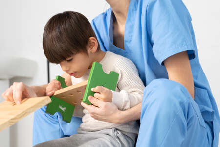 cute kid with disability playing with developing toys while is being helped by physiotherapist in rehabilitation hospital.の写真素材