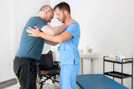 Male Physiotherapist helping a patient with a disability who uses a wheelchair, to get up at rehabilitation hospital.の写真素材