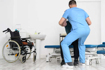 Male Physiotherapist helping a patient with a disability who uses a wheelchair, to get up at rehabilitation hospital.の写真素材