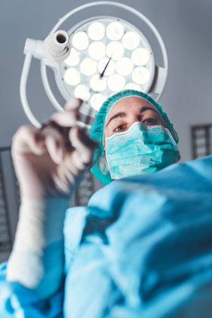 From below woman surgeon in medical uniform using professional tools while standing under bright...
