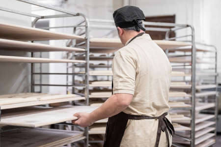 Baker placing tray with formed raw dough on rack trolley ready to bake in the ovenの写真素材