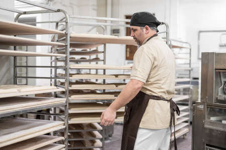 Baker placing tray with formed raw dough on rack trolley ready to bake in the ovenの写真素材