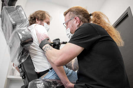Tattooist with his machine tattooing on the arm with the design drawn and using protective mask against covid 19, concept of art and design during the new normal.の写真素材