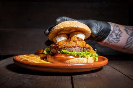 Close up of an unrecognizable chef finishing up a Premium quality Beef burger with goat cheese and caramelized onions, served with French fries on a rustic plate over wooden tableの写真素材