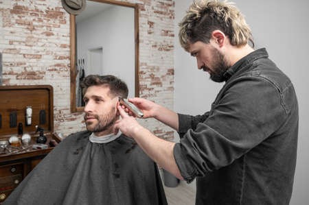 Man barber cutting hair of male client with clipper at barber shop. Hairstyling process.の写真素材