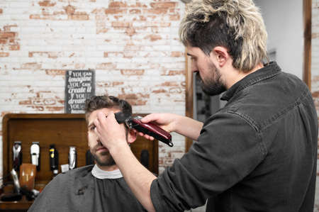 Man barber cutting hair of male client with clipper at barber shop. Hairstyling process.の写真素材
