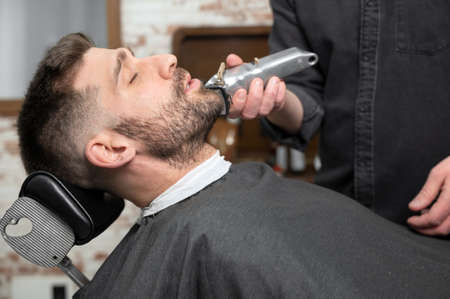 Young hipster Caucasian man during beard grooming in modern barber shop. Mens hair styling. Handsome man getting new hairstyle with electric trimmer.の写真素材