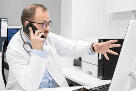 Male doctor pointing with finger on desktop computer while talking on the phone, discussing treatment with colleague.の写真素材