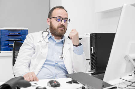 Portrait of friendly smiling healthcare professional therapist sitting at workplace. Happy confident male doctor physician wearing white medical coat and stethoscope.の写真素材
