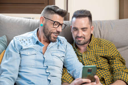 Happy young gay couple using mobile phone while sitting on a sofa in the living roomの写真素材