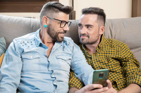 Cheerful gay couple sitting on the couch hugging and looking at the mobile phone, watching a movie, scrolling through the social networkの写真素材