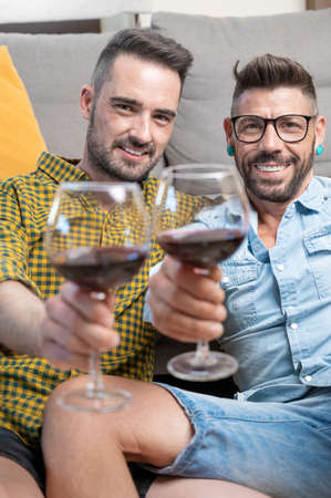 Gay couple in love sitting at home on the floor and toasting with red wine.の写真素材