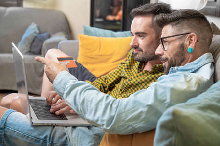 Happy young gay couple using laptop computer while sitting on a couch at home, shopping online with a credit cardの写真素材