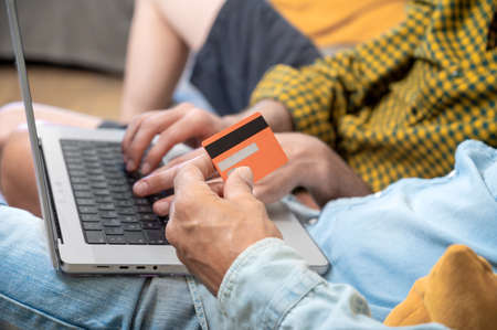 Close up of an unrecognizable gay couple using laptop computer while sitting on a couch at home,...