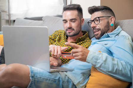 Happy young gay couple using laptop computer while sitting on a couch at home, shopping online with a credit cardの写真素材