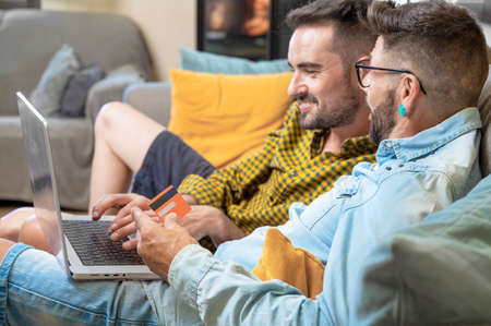 Happy young gay couple using laptop computer while sitting on a couch at home, shopping online with a credit cardの写真素材