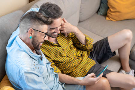 Cheerful gay couple sitting on the couch hugging and looking at the mobile phone, watching a movie, scrolling through the social networkの写真素材