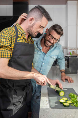 Happy hipster gay couple in love standing in the kitchen. They are cooking dinner together at home.の写真素材