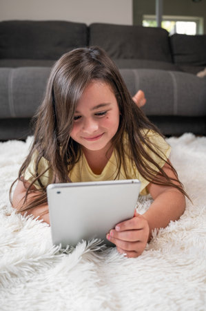Cute little girl watch cartoons on digital tablet. Kid lies on floor laughing using electronic device. Indoor leisure for children.の写真素材