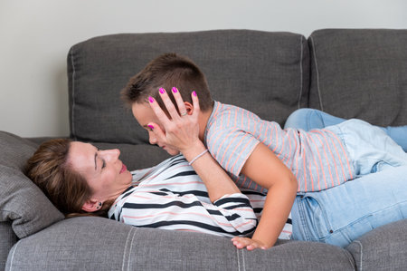 Woman lying on sofa kissing her little son in cheek. Mom with little boy joyfully spending time in cozy living room at homeの写真素材
