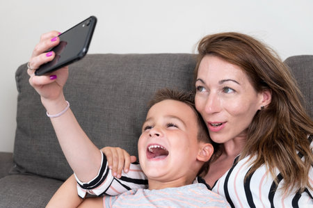 Cheerful mother holding smart phone making selfie with her little son showing victory sign. mom and kid boy sit on couch using gadget having fun taking self-portrait spend time together at home .の写真素材