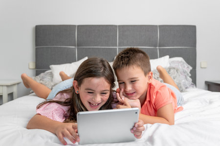 Cute little children with digital tablet using internet at home. Caucasian little sister and brother siblings surfing Internet, watching animated cartoons online on the bed.の写真素材