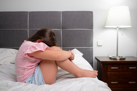 Girl crying in tears sitting on bed in bedroom. Unhappy children feeling sad lonely and upset in dark room at night at home.の写真素材
