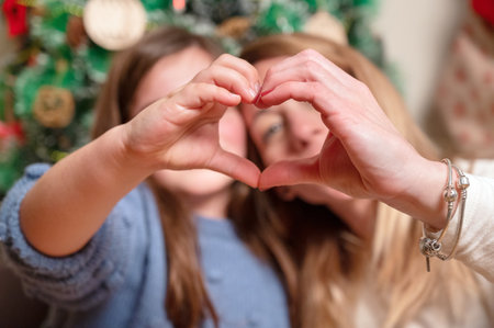 Loving family spends winter holidays at home. Portrait of happy smiling beautiful mother and child joining hands making heart shape sitting in festive, cozy room with Christmas tree.の写真素材