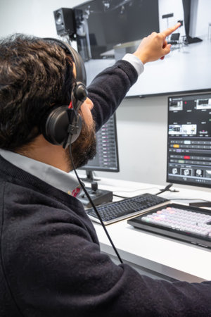 A television director in a broadcast control room, actively guiding a live production. Surrounded by multiple screens, the director wears a headset and gestures towards a video switcherの写真素材