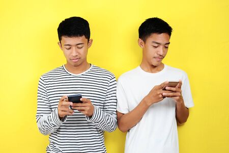 Portrait of handsome two young Asian men looking at their phone, isolated on yellow backgroundの写真素材