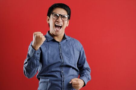 Young asian man happy and excited expressing winning gesture. Successful and celebrating on red backgroundの写真素材