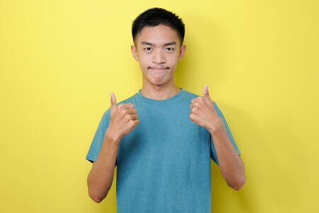 Young Asian man close his mouth giving two thumbs up, isolated on yellow backgroundの写真素材
