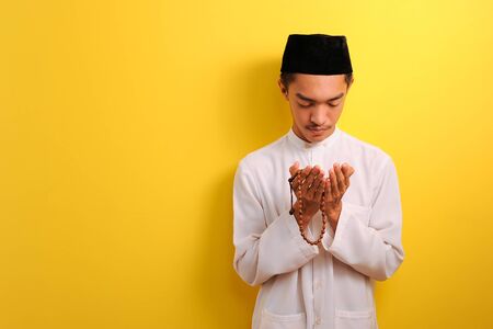 Young Asian Muslim man praying raising his hand with holding prayer beads, isolated on yellow...