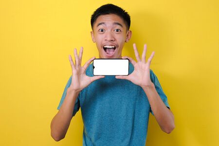 Happy Excited Young Asian man shock showing white phone screen at camera, isolated on yellow...