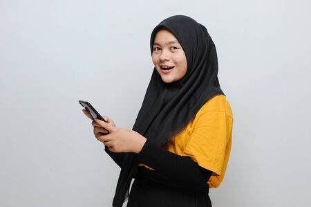 Portrait of a beautiful young Asian Muslim woman holding a smartphone look at a camera, isolated on grayの写真素材