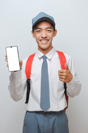 High School Student with uniform look at teh camera to showing white phone screen display to advertise something with thumb up, isolated on grey backgroundの写真素材