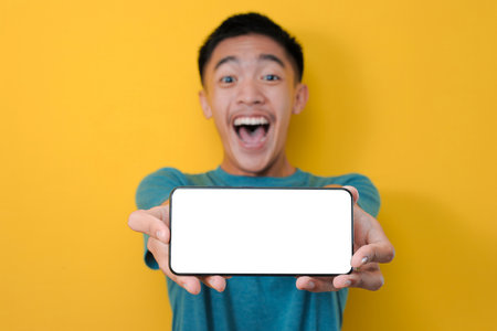 Happy Excited Young Asian man shock showing white phone screen at camera, isolated on yellow...