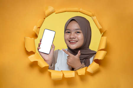 Young Asian teenager girl poses through torn yellow paper hole, showing mobile phone with blank white screen display for a copy space, wearing purple t-shirt and hat. Yellow isolatedの写真素材