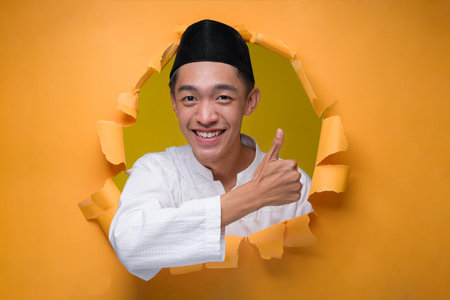 Asian Muslim Teenager man smiling at the camera with thumb up to copy sapce, poses through torn...