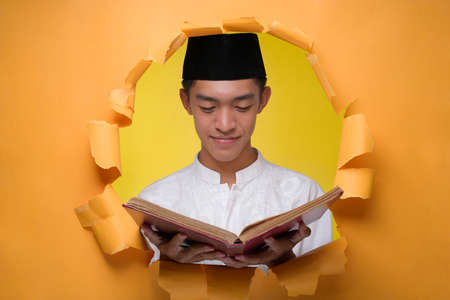 Happy Asian muslim man holding the Holy Quran, wearing muslim cloth with skull cap, poses through torn yellow paper hole, Reading The Holy Quran at Ramadan Kareemの写真素材