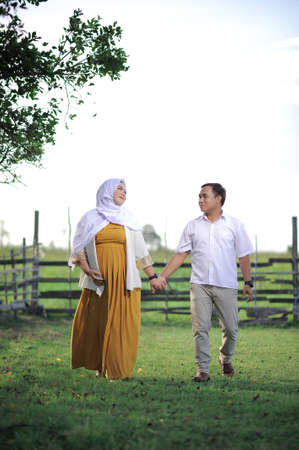 Affectionate young Asian Muslim couple in love wait for child, walk and hold together on green grass, have positive expression, want to become parents soon. Maternity, fatherhood and pregnancy.の写真素材