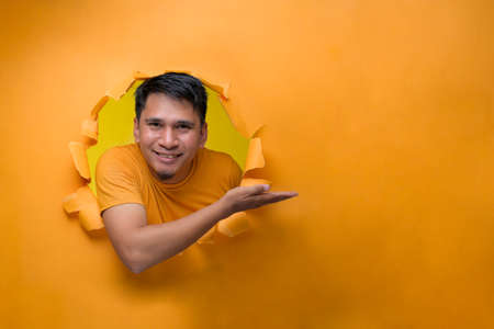 Young smiling Asian man through torn yellow paper hole showing a copy space to advertise a product. Advertising and Businessの写真素材