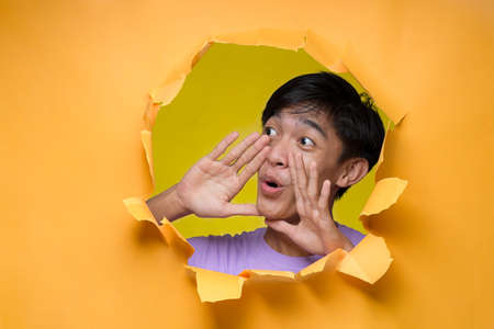 Young Asian Man through a hole paper making yelling gesture