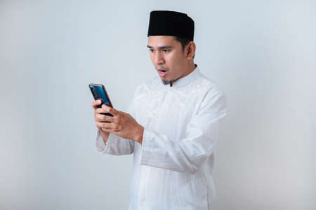 Shocked Muslim man wearing muslim clothes holding mobile phone look at phone screen against on white backgroundの写真素材
