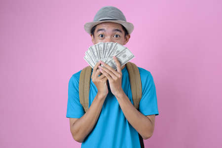 Portrait of a joyful young asian traveler man holding some money with backpack and hat, smiling against on pink backgroundの写真素材