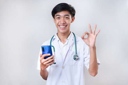 Happy Young Asian doctor smiling showing okay sign holding a mobile phone, against on white backgroundの写真素材