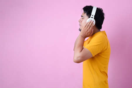 Asian man in casual yellow t-shirt wearing headphones listening to music, isolated on pink background.の写真素材